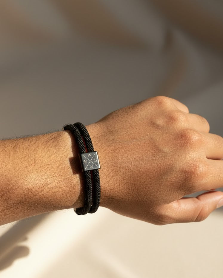 Hand wearing a black braided bracelet with a square metal clasp on a neutral background